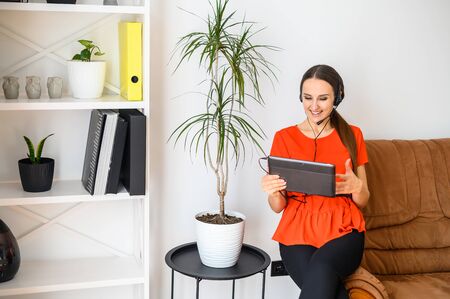 Morning meeting via video, video conference from home. Young woman sit on the edge of sofa with a tablet, she using headset for online video conversationの写真素材