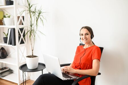 Stay at home. Work at home, distant work. A young woman sits on a chair using hands free headset and laptop for remote workの写真素材