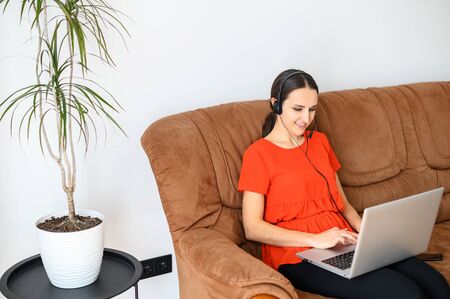 Young woman work from home. She is sitting on sofa with a laptop and using headset for online talks. Concept of remote workの写真素材