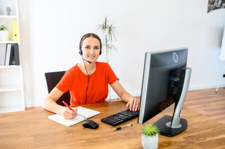 Business woman working online, using headset and personal computer. She writes something in notebook, looks at camera and smilesの写真素材