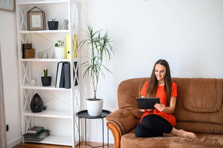 A young woman spends her leisure time surfing the web, watching movies, at social networks. She is sitting on a sofa with a tablet, looking at the screen and smiling.の写真素材