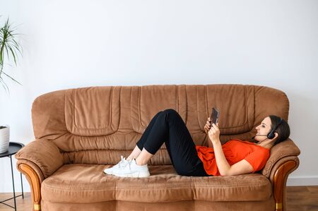 Video meeting, video chat. Young woman with hands free headset and tablet lies on a sofa and looks at the screenの写真素材