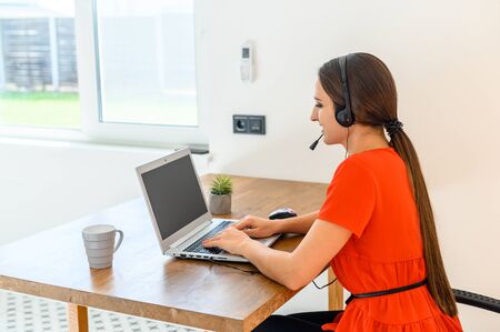 Remote work, office at home, work from home. A young woman uses a headset for an online talks, she sits at a table with a laptop. View from the backの写真素材