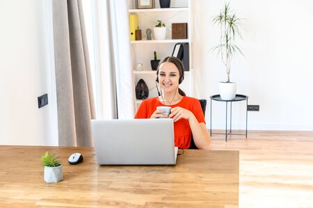 Young woman call center operator, support service operator at home office. A woman uses a headset to talk, she sits with a cup of coffee at a table, looks at a laptop screen and smilesの写真素材