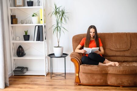 Stay home. Home leisure during quarantine, epidemic. A young woman is reading a book, she is sitting on the sofa. Cozy home atmosphereの写真素材
