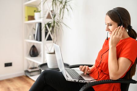 Stay at home. Work at home, distant work. A young woman sits on a chair using hands free headset and laptop for remote workの写真素材