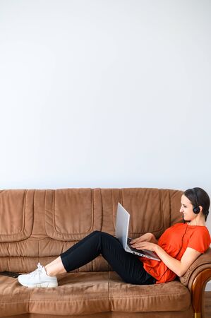 Remote work in a relaxed style. A young woman works from home using hands free headset and laptop, she lies on the sofa, talking and looking at the screenの写真素材