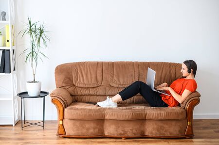 Remote work in a relaxed style. A young woman works from home using hands free headset and laptop, she lies on the sofa, talking and looking at the screenの写真素材