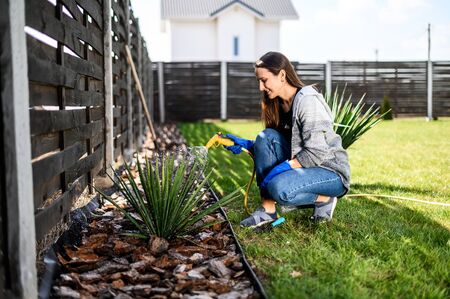 The concept of spring gardening, a hobby. Young positive woman in gloves with hose is watering a plants, he crouched on green grass. Gardening at backyardの写真素材