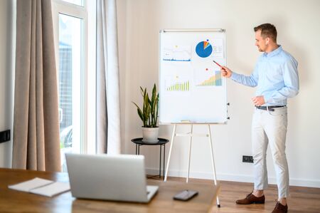 Webinar, online meeting with colleagues. Young attractive businessman in smart casual wear makes online presentation. He stands near the flip chart and points to it with a pen, laptop in front of himの写真素材