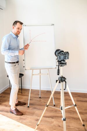 Online tutor, teacher, mentor. Positive guy in formal shirt stands with a marker in hand near white board, looks at camera on tripod and smiles. He is giving a video lesson. Concept of online studyingの写真素材