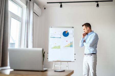 Concentrated bearded guy in a formal shirt looks at a graphs and diagrams on whiteboard, he solves business tasks, laptop in front of him. Concept of webinars, online meetingの写真素材