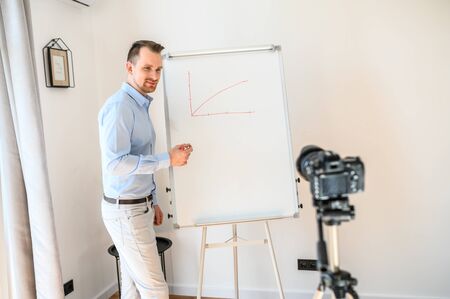 Online tutor, teacher, mentor. Positive guy in formal shirt stands with a marker in hand near white board, looks at camera on tripod and smiles. He is giving a video lesson. Concept of online studyingの写真素材