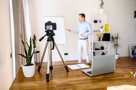 Business coach, mentor. A young guy is recording business vlogs, he is standing near a flip chart with his arms crossed on his chest in a confident pose, a camera on the tripod and a laptop on the table.の写真素材