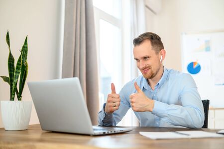 Video communication, video call using laptop. A young guy an office worker in a formal shirt with airpods in his ears sits at a table and speaks on video communication. He smiles and shows thumbs upの写真素材