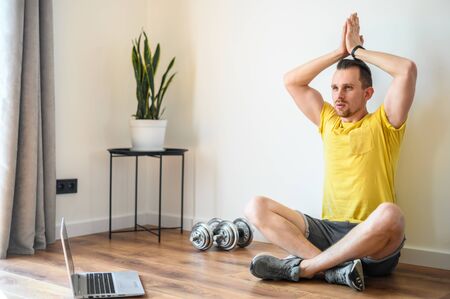 The concept of online training, sports video lessons. Young athletic guy practices yoga at home with online lessons. Laptop in front of himの写真素材