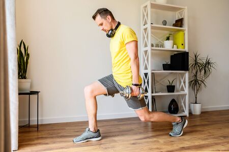 Do yourself better. Young active guy doing lower body workout at home. He doing lunges with dumbbells. The concept of a healthy lifestyle, self-discipline, sporty styleの写真素材