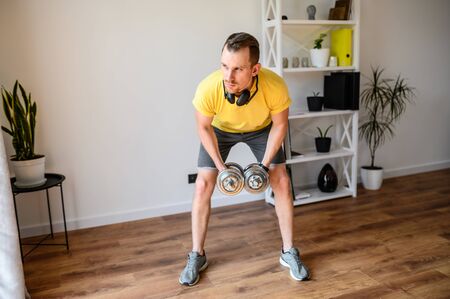 Stay home. A guy has upper body workout at home. He is doing exercises for arms and shoulders with iron dumbbellsの写真素材