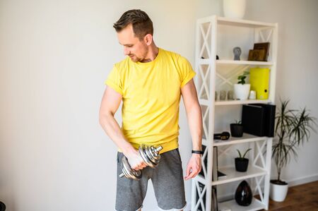 Upper body workout at home. Athletic guy shakes his biceps using iron dumbbells.の写真素材