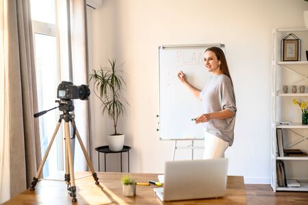 Concept of webinars, online classes, online training. Young woman in casual clothes shows information on a flipchart board and records herself on camera on tripodの写真素材