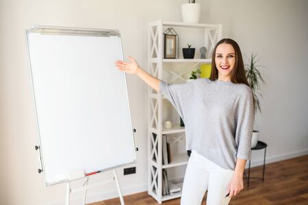 Tutor, mentor, teacher. Young attractive woman stands and points on flipchart board and smiles indoorsの写真素材