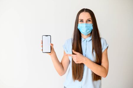 World pandemic, epidemic. Young woman in medical mask with a smartphone in her hand, she points a finger at the screen. Isolated on white backgroundの写真素材