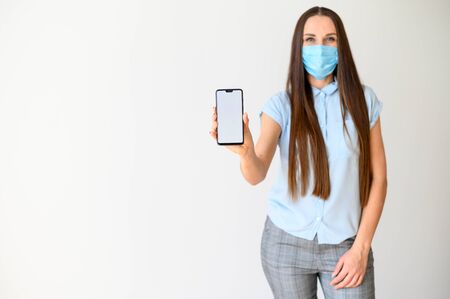 Worldwide pandemic, epidemic. Young woman in medical mask with a smartphone in her hand, she demonstrates the screen of the phone at the camera. Isolated on white backgroundの写真素材