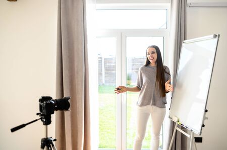 Young woman record tutorial video how to learn foreign language online. She stand indoors with a flipchart near, writes on it and explains something, camera recording video. Online Learning Conceptの写真素材