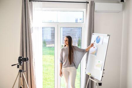 Young woman is recording a video presentation, video lesson indoors. She stands and shows the pen on a flipchart with graphs and charts and looks in camera on tripodの写真素材