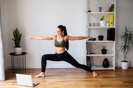 Yoga video tutorials. Girl training at home, practicing yoga and watching videos on laptop in living room. She is doing warrior asana. Healthy lifestyle conceptの写真素材