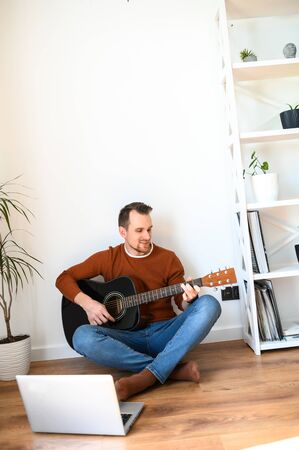 Hobbies and leisure during quarantine, on self-isolation. Attractive guy watching guitar video tutorials. Homely cozy atmosphere, a guy sits on a floor with an acoustic guitar in his handsの写真素材
