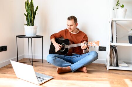 Online learning. Guy watches guitar lessons videos on laptop and repeats while sitting on the living room floorの写真素材