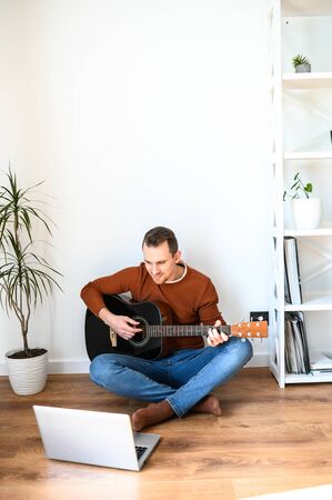 Online training, online classes. A guy watches video tutorial on guitar playing, he sits on a cozy living room with a guitarの写真素材