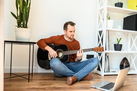 Online training, online classes. A guy watches video tutorial on guitar playing, he sits on a cozy living room with a guitarの写真素材