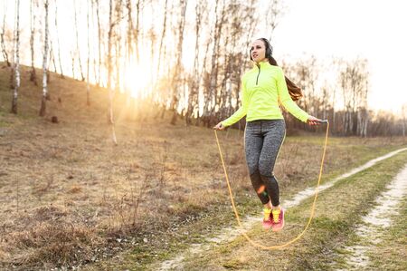 Training with skipping rope. Woman with headphones in neon green sportswear is jumping outdoorsの写真素材