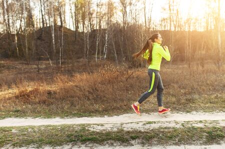 Woman runner in motion. A young athletic woman jogging in the countryside, she in bright athletic uniform runs on a dirt road in the forestの写真素材