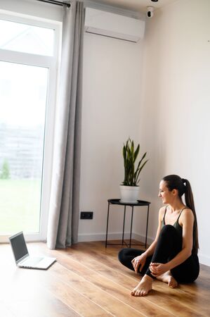 Yoga for beginners. A young woman in sportswear is using a laptop to watching yoga video tutorial at home. She sits on the floor, looks at the screen. Sport at home conceptの写真素材