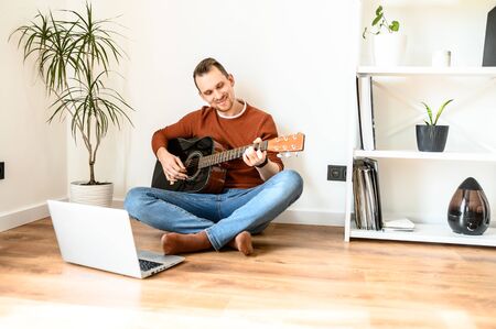 Online training, online classes. A guy watches video tutorial on guitar playing, he sits on a cozy living room with a guitarの写真素材