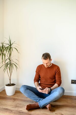 Young guy chatting on the phone. He sits on the floor in the living room of the house and types on the screen of the smartphone and smiles.の写真素材