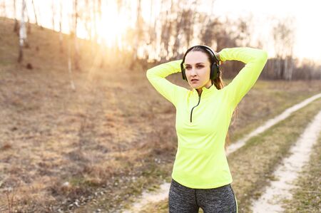 Young attractive woman in trendy sportswear with a headphones ties her hair in a bun before doing workout, she is going to run outdoors. Sport lifestyleの写真素材