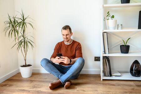 Young guy chatting on the phone. He sits on the floor in the living room of the house and types on the screen of the smartphone and smiles.の写真素材