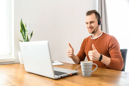 Work from home, home office. A young guy in casual clothes is using a headset for online communication. He sits at the table, looks at the laptop screen, speaks and shows thumbs up. Everything okの写真素材