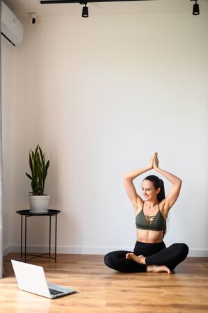 Young woman in sportswear is studying yoga online. She sits in the living room on the floor in a lotus position, looks at the laptop screen. Healthy lifestyle conceptの写真素材