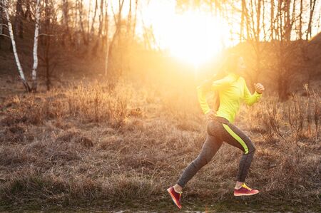 Woman runner in motion. A young athletic woman jogging in the countryside, she in bright athletic uniform runs on a dirt road in the forestの写真素材