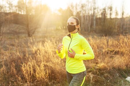 Athletic woman in bright sportswear and in protective medical mask outdoors at sunset. The concept of sport workout on quarantine period, epidemic of coronavirusの写真素材