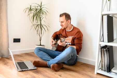 Online learning. Guy watches guitar lessons videos on laptop and repeats while sitting on the living room floorの写真素材