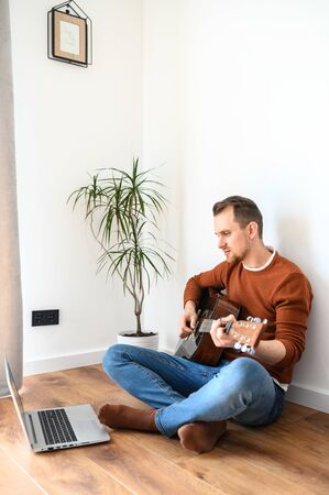 Online training, online classes. A guy watches video tutorial on guitar playing, he sits on a cozy living room with a guitarの写真素材