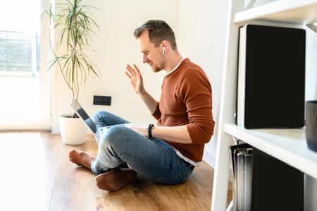 Self-isolation during quarantine. Video communication, zoom. Young positive guy in a casual wear communicates via video communication using a laptop, he waving hello to the screen. Side viewの写真素材