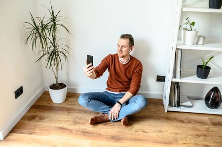 The guy takes a selfie, speaks via video. A young guy sits at home on the floor in the living room, takes pictures of himself on the front camera of the phone and smilesの写真素材