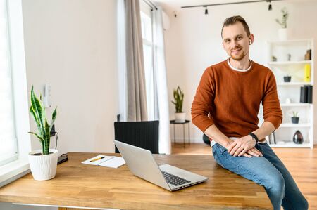 Home office. A young businessman uses laptop for work, he sits on the edge of the table, looks to the camera and smiles.の写真素材
